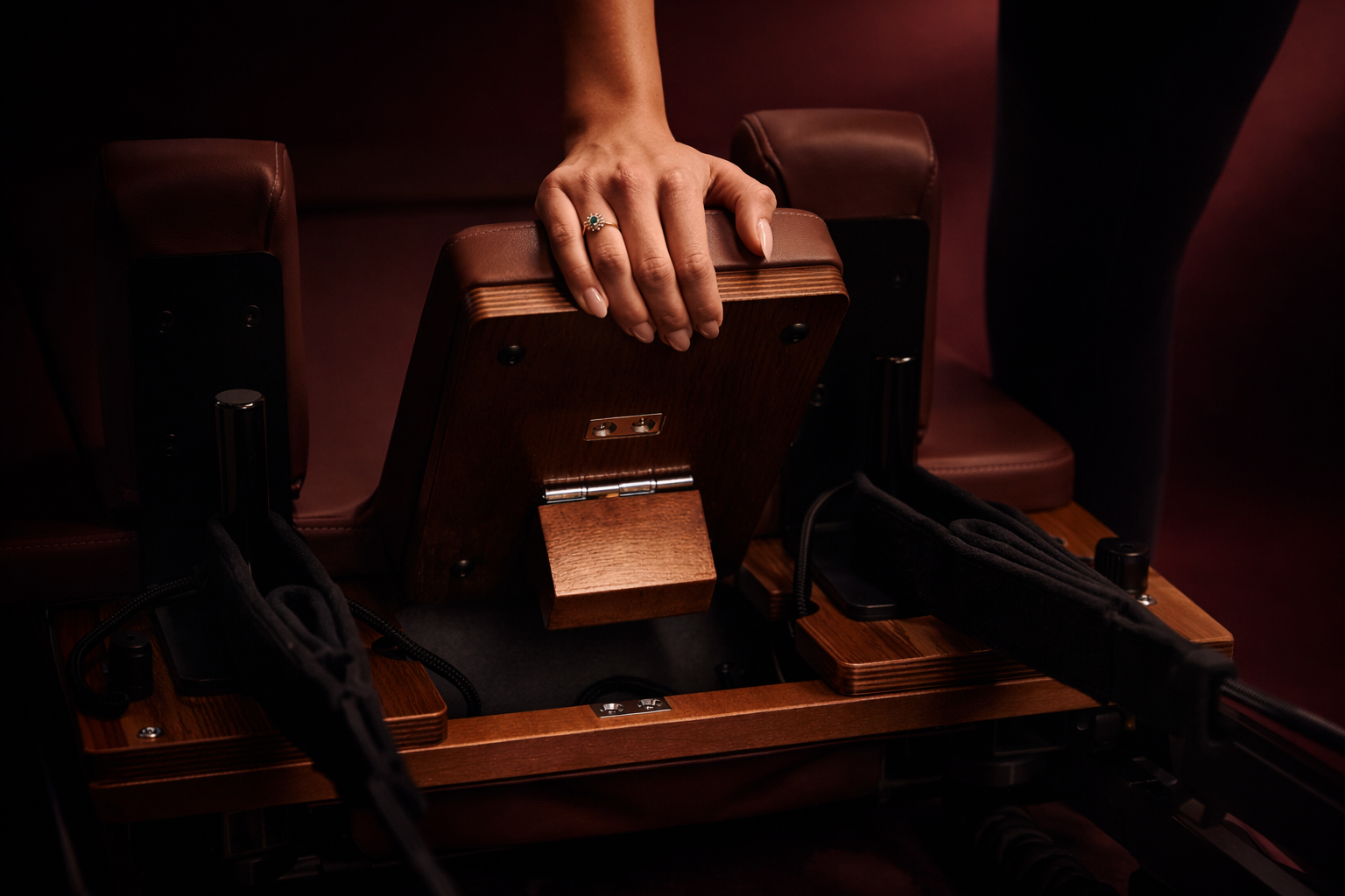 Close-up of a white woman's hand adjusting a wooden reformer Pilates footboard in a softly lit, burgundy-toned studio