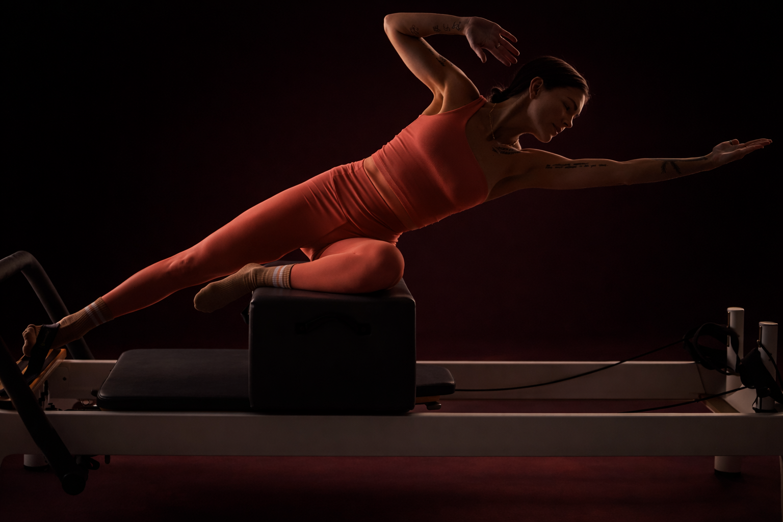 Female form on a Reformer Pilates bed striking a Side Stretch Pilates power pose against deep burgundy backdrop