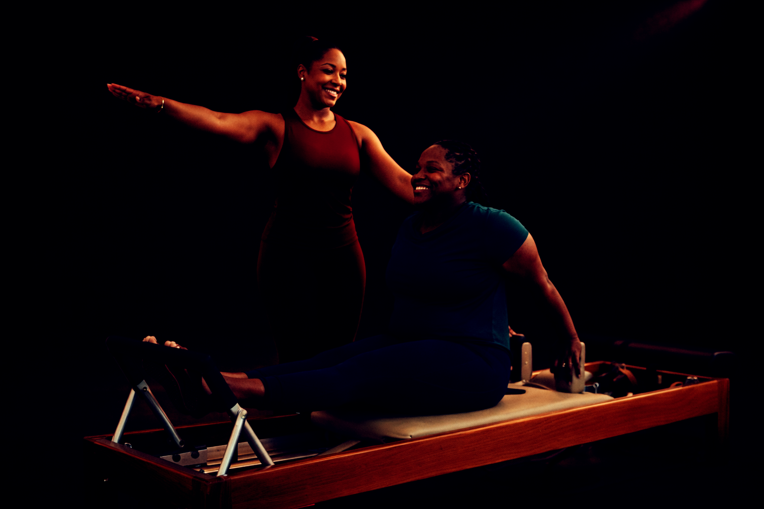 Pilates session in moody lighting doing a 121 session with instructor instructing a large black lady on a reformer chicken