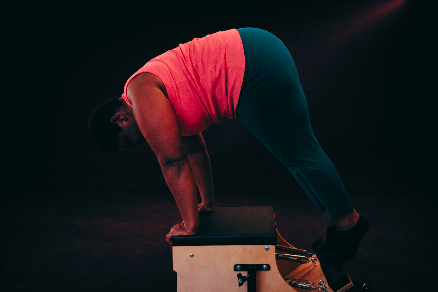 Large build Woman performing a plank on a reformer Pilates machine in a dark, burgundy-toned studio with soft directional lighting wearing a coral gym top and teal leggings