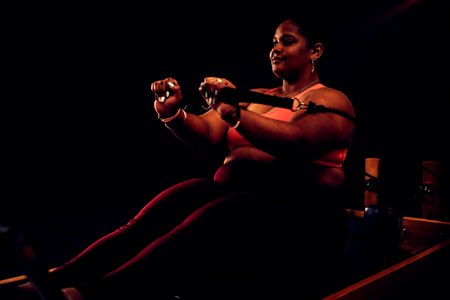 Large black lady sitting on a reformer bed pulling the short straps