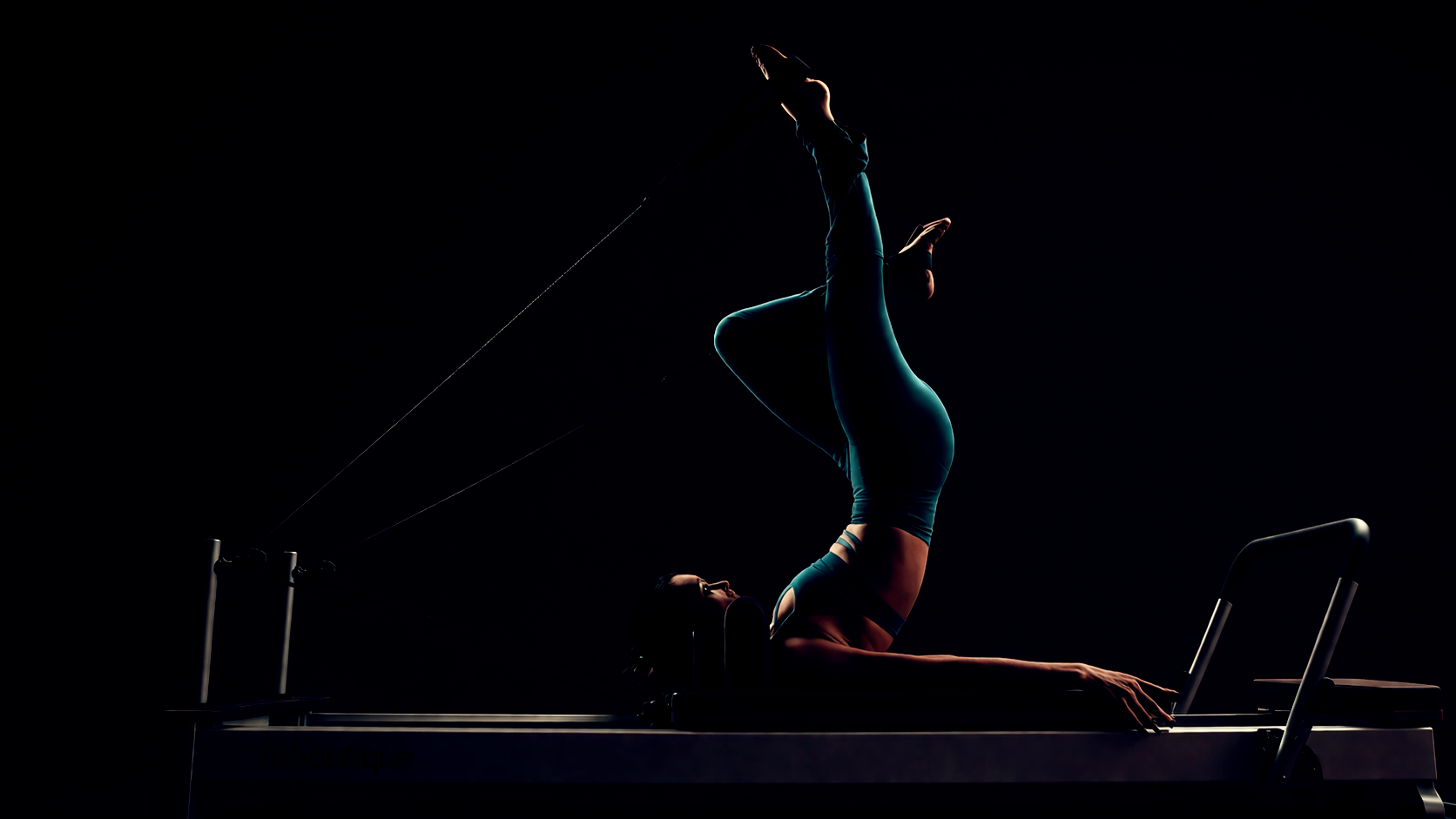 Woman performing an inverted leg exercise on a reformer Pilates machine in a dark burgundy studio with soft directional lighting wearing a teal gym kit